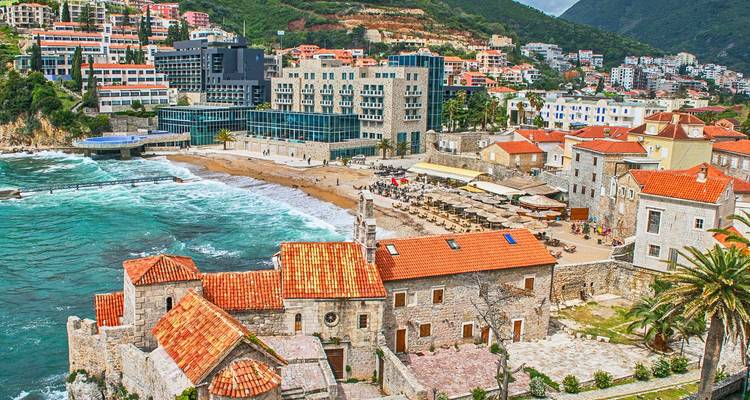 Seaside view of Budva with stone church, sandy beach, sun-loungers and modern hotels rising up the green hillside