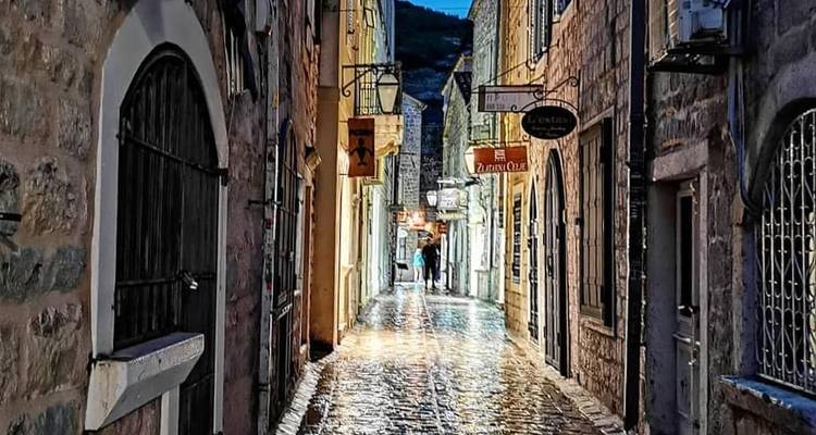 Narrow cobbled medieval alley at dusk, lit by warm lanterns with shop signs hanging overhead and a lone walker in the distance