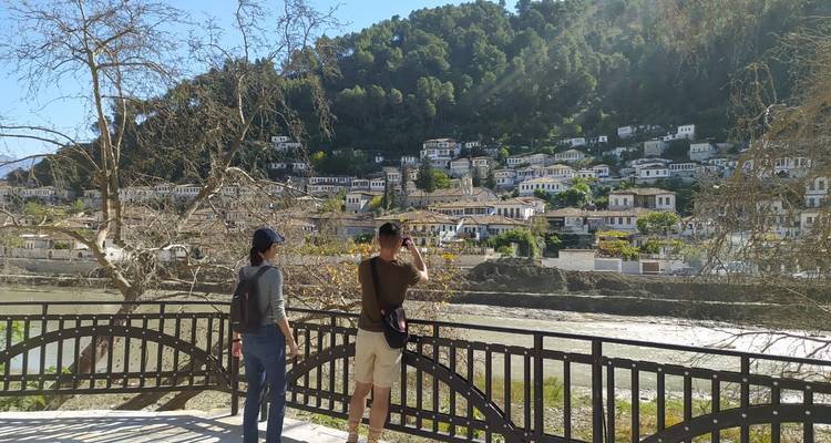 Two travellers look across a river toward the terraced white houses of Berat spread up a forested hillside