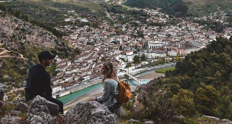 Couple sitting on rocks high above Berat with a sweeping view of the river, bridge and red-roofed city below