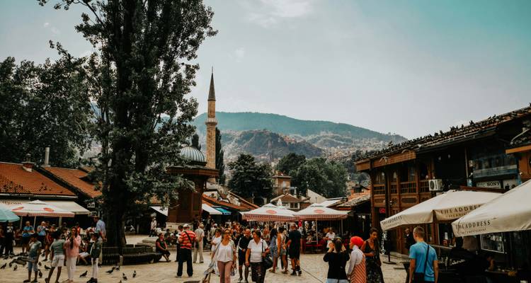 Plaza de bazar bulliciosa en Sarajevo con un minarete y colinas boscosas de fondo
