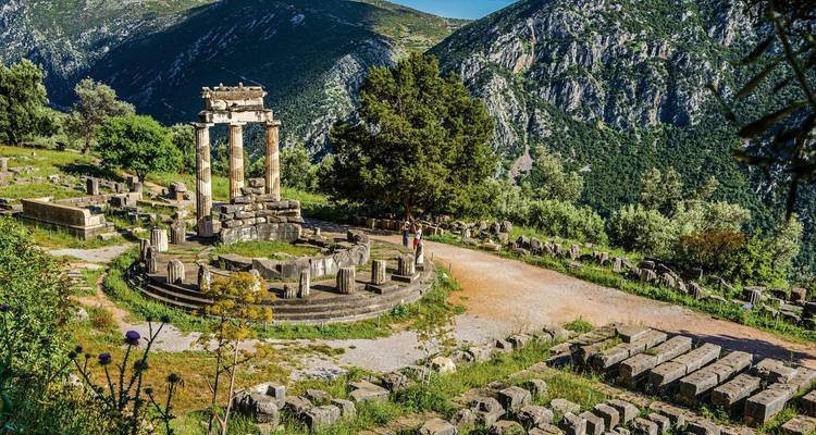 Ruinas del Tholos circular de Delfos enmarcadas por montañas verdes y escarpadas bajo cielos despejados