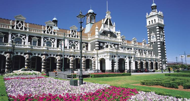 Façade ornée de la gare ferroviaire de Dunedin avec des parterres de fleurs colorés sous un ciel bleu profond