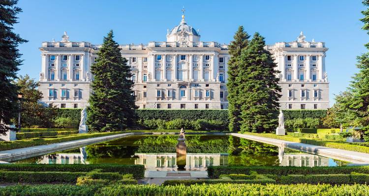 Formale Gärten spiegeln die prächtige Fassade des Königspalastes von Madrid wider, die von hohen Immergrünen unter klarem blauen Himmel gerahmt wird.