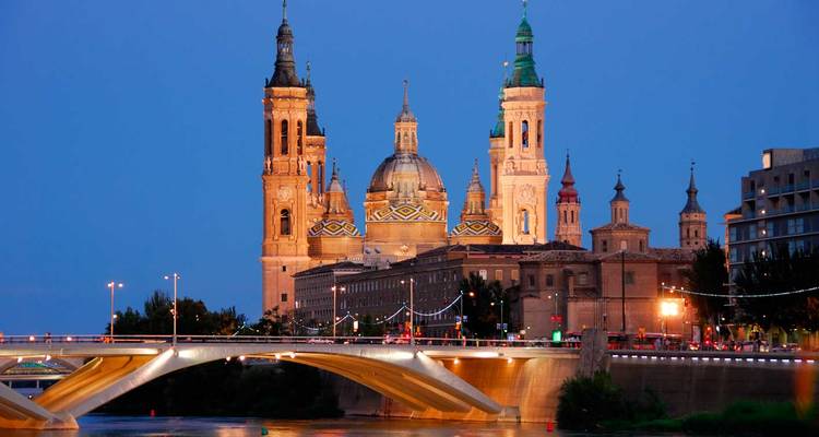 Basilika Unserer Lieben Frau del Pilar, beleuchtet zur blauen Stunde über dem Ebro und einer modernen Brücke in Saragossa.
