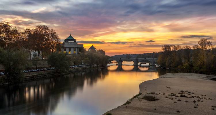 Serene sunset over a broad river reflecting fiery skies, with an arched stone bridge and riverside buildings lining the banks.