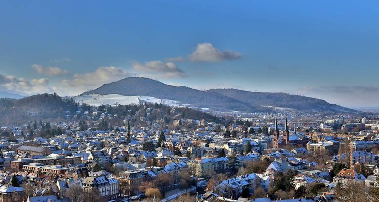 Wintry panorama of Freiburg with snow-topped roofs and the Black Forest foothills beneath a clear blue sky.