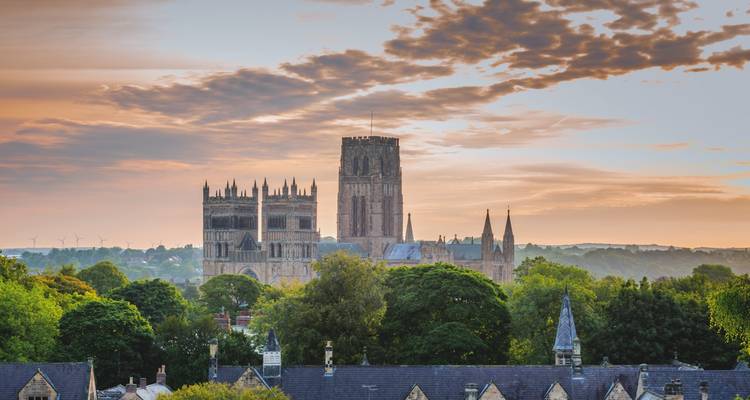 Durham Cathedral’s towers rise above treetops under a colorful sunset sky.