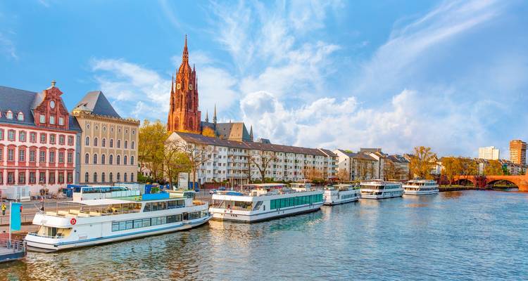 Sunny riverside scene in Frankfurt with cathedral spire and cruise boats lined along the Main River.