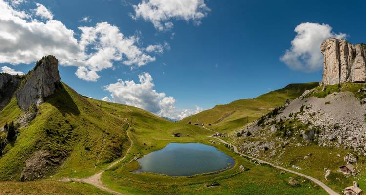 Prado alpino con un pequeño lago circular, picos escarpados y senderos para caminar bajo un cielo azul