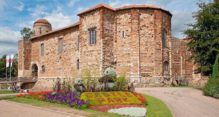 Château normand construit en pierre rougeâtre entouré de parterres de fleurs colorés dans un parc