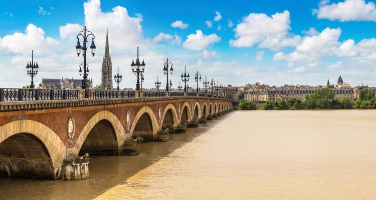 Le Pont de Pierre, pont de pierre enjambant le fleuve Garonne avec l'horizon de Bordeaux et sa flèche en arrière-plan sous un ciel partiellement nuageux.