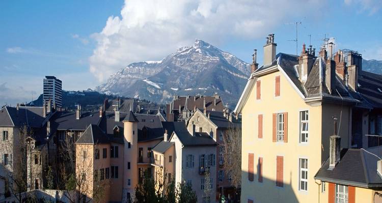 Rooftops of Chambéry with a snow-capped mountain rising behind under scattered clouds.