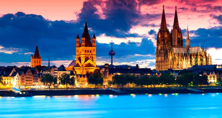 Dramatic sunset behind Cologne Cathedral and historic riverside skyline glowing over the Rhine.