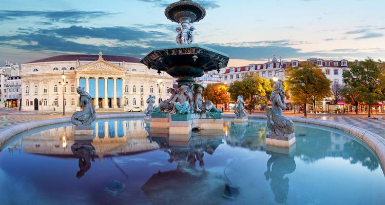Elegant fountain reflecting classical statues and the neoclassical National Theatre on Lisbon’s Rossio Square at sunrise.
