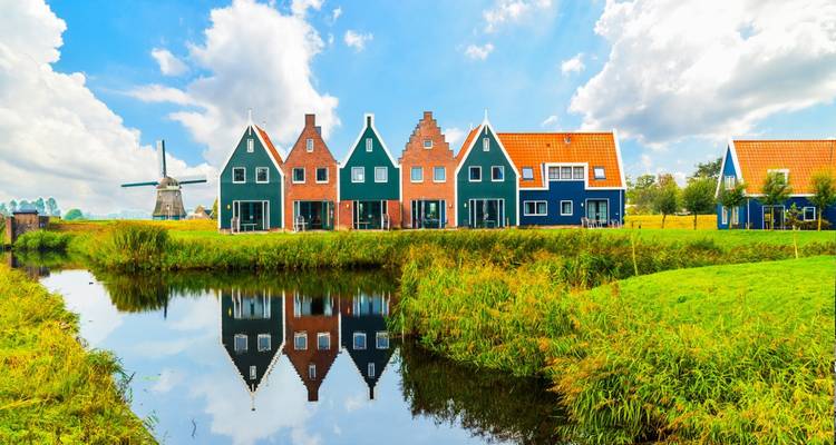 Des maisons traditionnelles colorées et un moulin lointain se reflétant dans un canal calme en milieu rural aux Pays-Bas