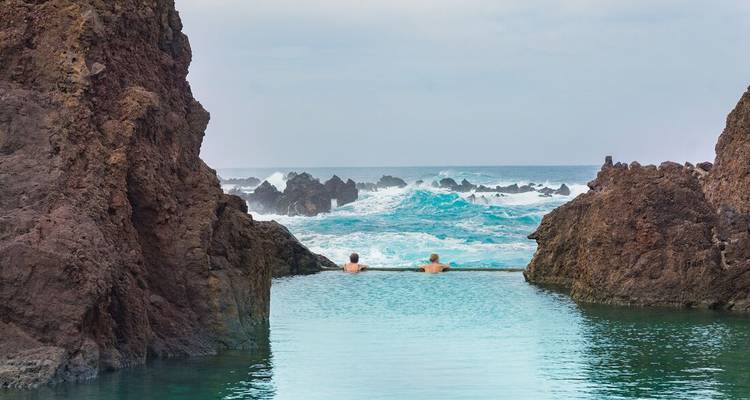 Pair of swimmers relax in a natural lava-rock ocean pool with waves crashing beyond.