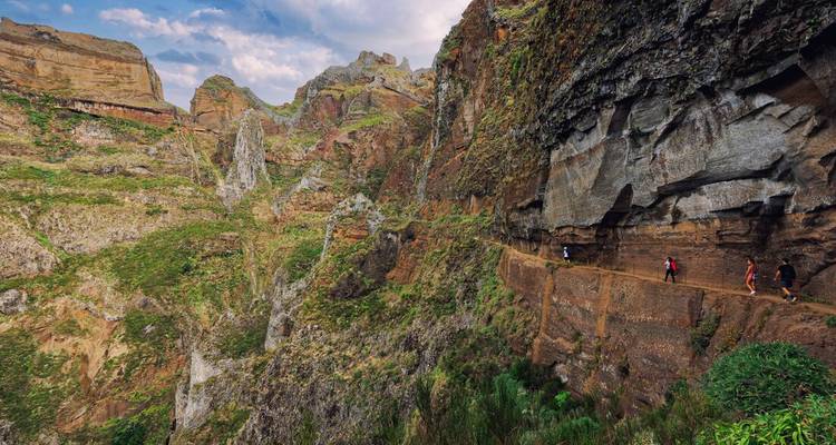 Hikers walk along a dramatic cliff-side levada path carved into towering green rock walls.