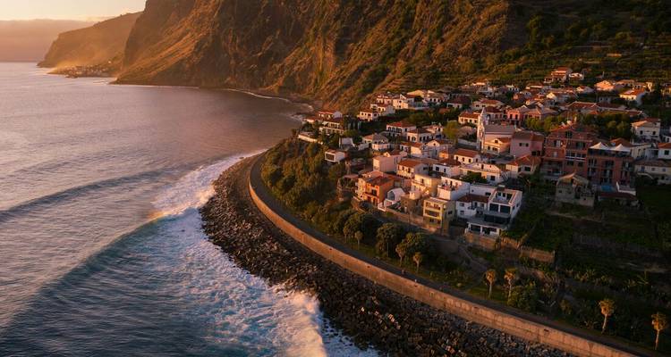 Golden-hour aerial of a seaside Madeira village hugging steep green cliffs above gentle waves.
