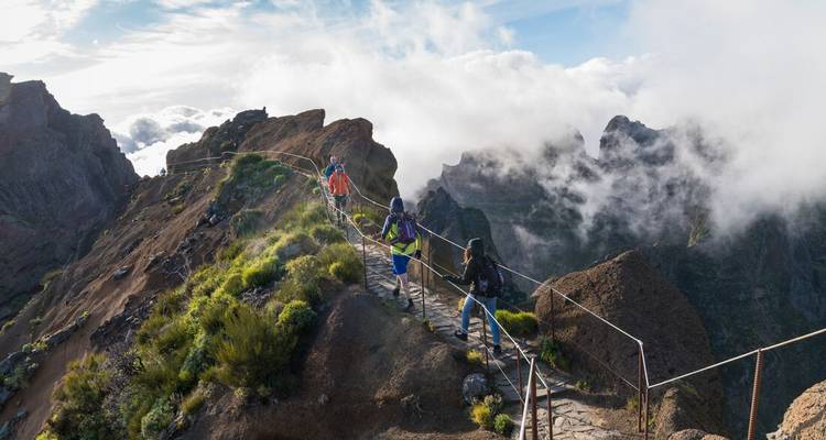 Group of trekkers traverse a narrow stone path above clouds on a rugged mountain ridge.