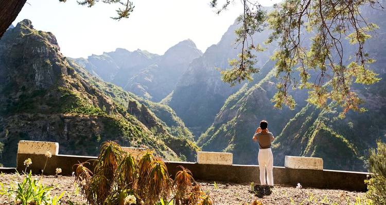 Traveler photographs a lush green mountain valley from a shaded roadside viewpoint.