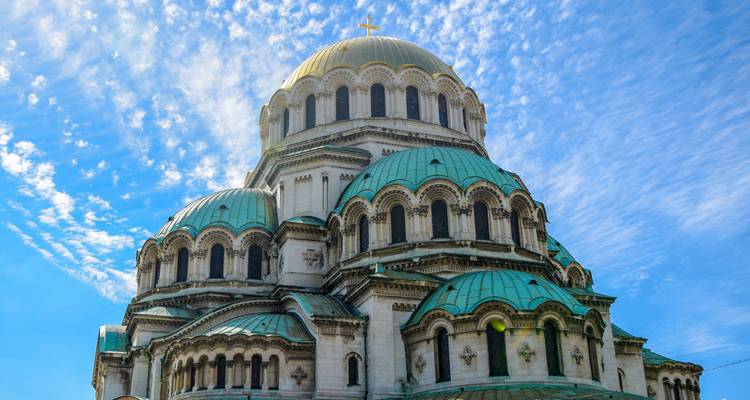 The grand Alexander Nevsky Cathedral with its green domes gleams beneath a vibrant blue, partly-cloudy sky.