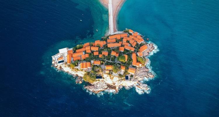 A stunning aerial shot of the tiny red-roofed island village of Sveti Stefan connected to the mainland by a narrow causeway amid deep-blue Adriatic waters.