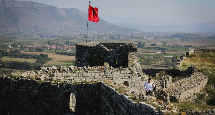 Stone ramparts of a hilltop Albanian fortress fly a vivid red flag while a lone visitor explores with sweeping valley views beyond.