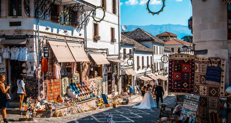 Colorful carpet-lined bazaar street on a steep stone lane bustling with shoppers beneath bright Balkan sunshine.