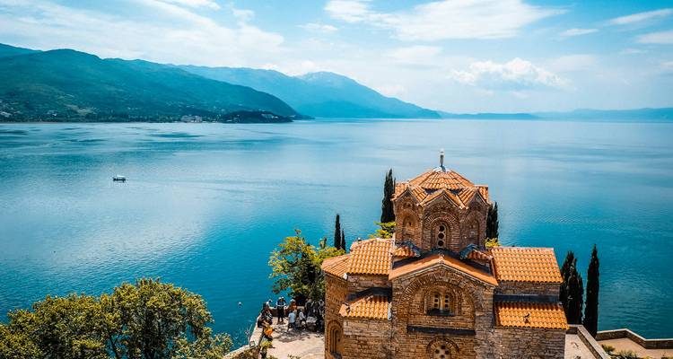 The lakeside Church of St John at Kaneo perches on a cliff above the tranquil blue waters of Lake Ohrid with distant mountains.