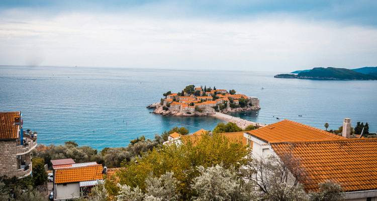Distant view of the picturesque islet of Sveti Stefan framed by coastal rooftops and calm Adriatic sea under a hazy sky.