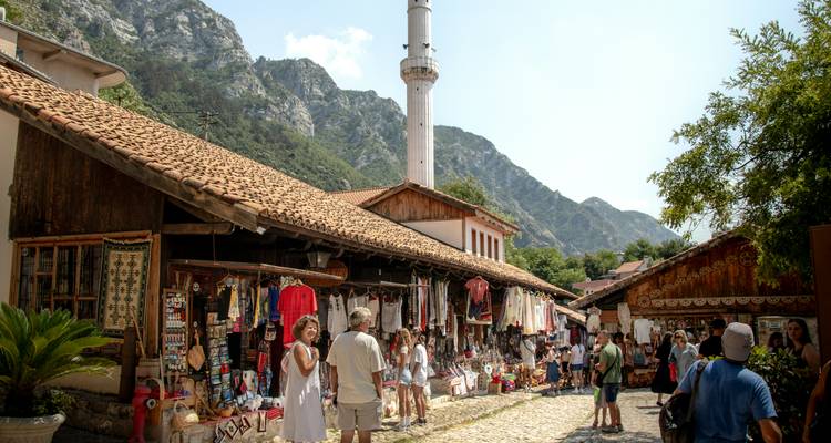 Mountain-backed village bazaar with minaret, rustic stalls and visitors browsing handicrafts along a cobblestone lane.