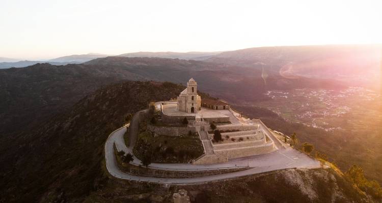 Prise de vue par drone d'un sanctuaire perché au sommet d'une colline et d'une route sinueuse baignés dans la lumière chaude du coucher de soleil sur les montagnes.