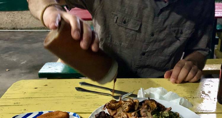 Gros plan d'un client versant de la sauce sur une assiette de poulet jerk à une table extérieure décontractée.