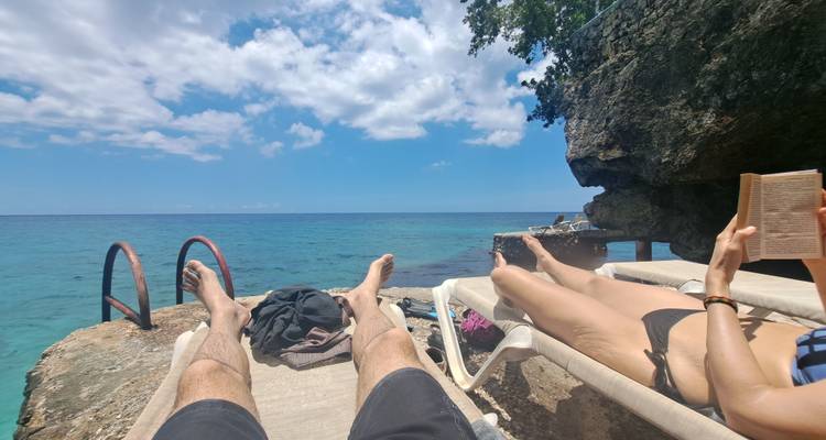 Deux voyageurs se détendent sur des chaises longues au bord de la mer à côté d'une eau turquoise, l'un lisant un livre avec un ciel bleu au-dessus.