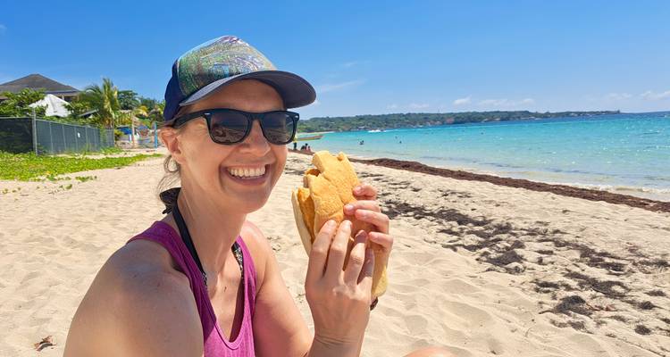 Femme souriante portant des lunettes de soleil dégustant un sandwich assise sur une plage des Caraïbes ensoleillée aux eaux turquoise.