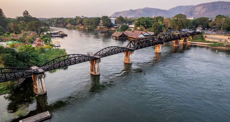 Aerial view of the historic iron Bridge over the River Kwai spanning a tranquil river with hills in the background.