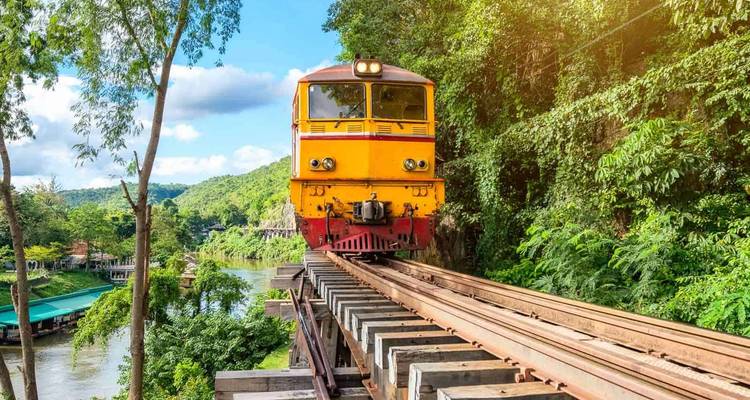 Bright yellow locomotive crossing a wooden trestle bridge through lush jungle over a river.