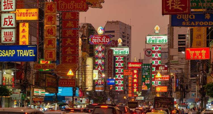 Busy Bangkok Chinatown street filled with neon Chinese signs glowing above traffic at dusk.