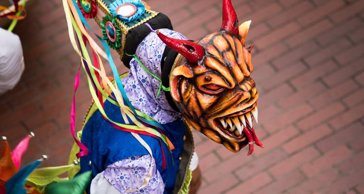 Levendige festivalbezoekersnaar met een fel duivelmasker en linten tijdens straatparade.