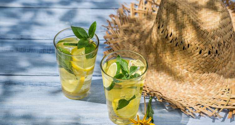 Verfrissende limonadeglazen met munt ernaast op een houten tafel, naast een stroien zonnehoed.