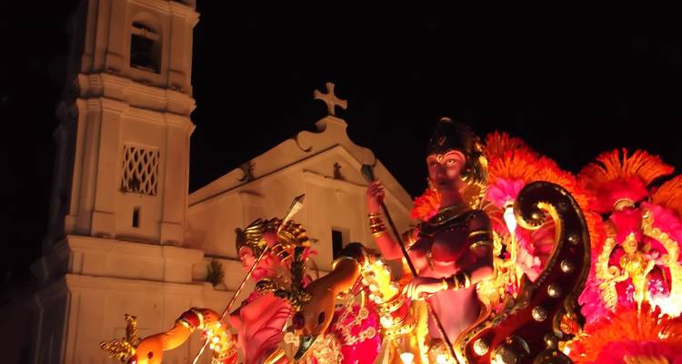 Verlichte carnavalwagen met mythologische figuren voor een koloniale kerk in de nacht.