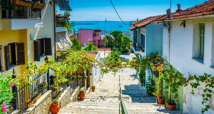 Callejón empedrado y empinado en el Ano Poli de Tesalónica lleno de casitas coloridas, viñas colgando arriba y vistas al mar a lo lejos.