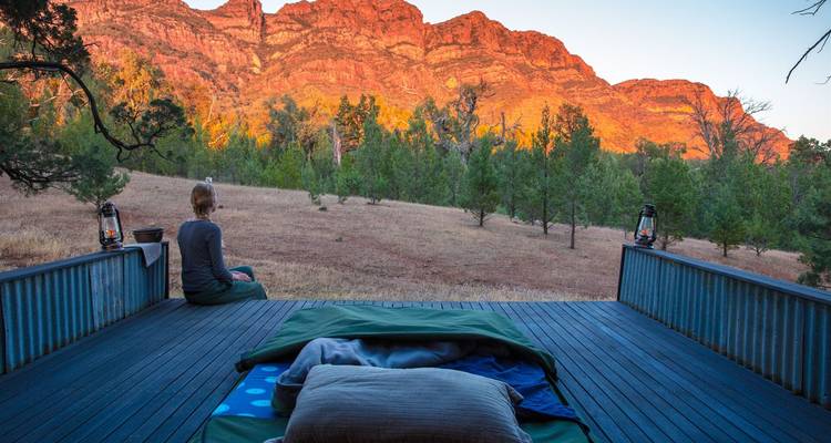 Ein Camper entspannt sich auf einer Veranda neben ausgerolltem Lagerausrüstung und blickt auf das in Sonnenlicht getauchte Wilpena Pound.