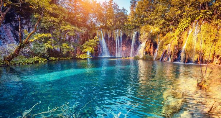 Türkisfarbener See mit kristallklarem Wasser, das kaskadenartige Wasserfälle inmitten eines üppigen Waldes bei goldenem Sonnenaufgang widerspiegelt.