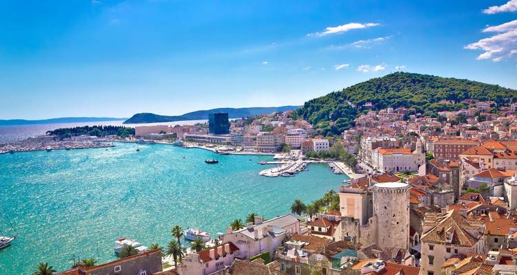 Lebendiges Küstenpanorama des Hafens von Split, türkisfarbenes Meer, historische Steingebäude und grüner Hügel.