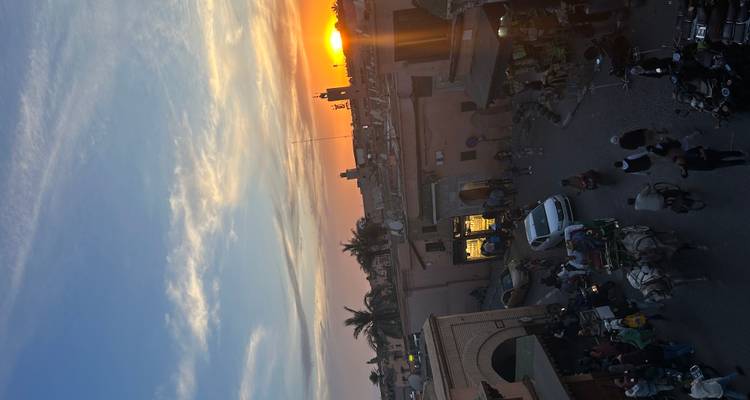 Coucher de soleil sur les terrasses de Marrakech avec une scène de rue animée et des calèches tirées par des chevaux.
