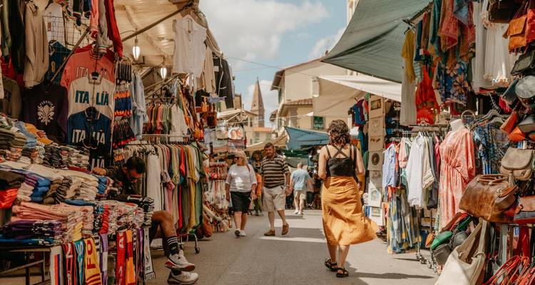 Callejón de mercado al aire libre lleno de vida con ropa y souvenirs, compradores paseando bajo los toldos.