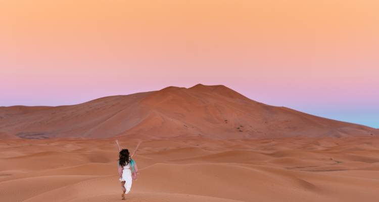 Ein einzelner Reisender spaziert bei Sonnenuntergang in Pastelltönen über sanfte orangefarbene Wüstendünen