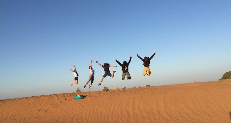 Five travellers mid-air jumping from a dune ridge against a clear sky.
